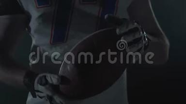 不可辨认的美国足球运动员在运动器材装备手中的球在黑暗的背景下紧紧地旋转着